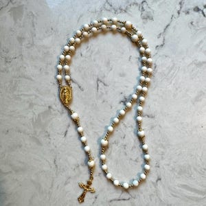 May include: A rosary featuring white beads interspersed with small gold-toned metal beads. The piece includes a gold-toned metal centerpiece depicting a figure and a gold-toned crucifix pendant at the bottom, connected by a delicate metal chain.