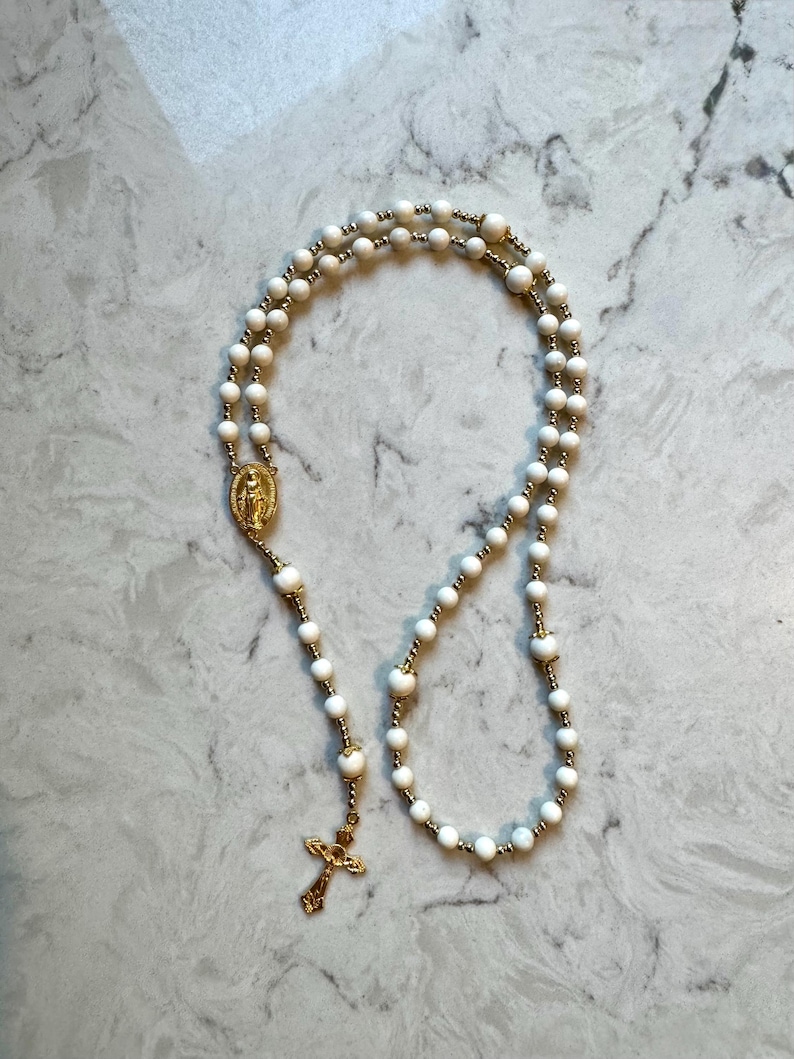 May include: A rosary featuring white beads interspersed with small gold-toned metal beads. The piece includes a gold-toned metal centerpiece depicting a figure and a gold-toned crucifix pendant at the bottom, connected by a delicate metal chain.