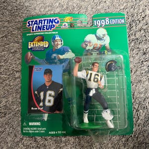 May include: A 1998 edition Starting Lineup sports collectible figure of a football player in a white and navy uniform, number 16. The packaging is green with the words "Starting Lineup" and "Extended Series". Includes a trading card of Ryan Leaf.