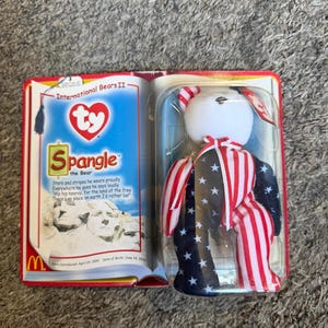 May include: A Spangle the Bear Ty Beanie Baby in its original packaging. The bear is red, white, and blue with stars and stripes. The packaging includes the text "International Bears II" and a poem.