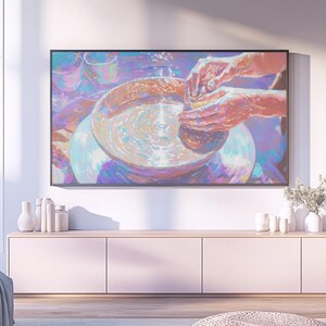 May include: A framed painting showing hands working clay on a pottery wheel. The artwork uses blues, purples, and oranges. The painting is above a light pink cabinet with white vases and decorative items.