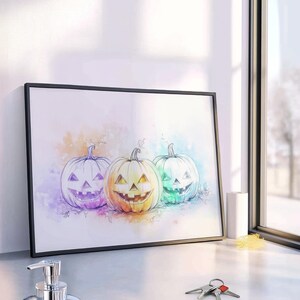 Könnte beinhalten: Ein gerahmter Druck mit drei lächelnden Kürbissen im Aquarellstil. Die Kürbisse sind in Lila, Gelb und Grün gehalten, mit schwarzen Umrissen und einem weißen Hintergrund. Das Kunstwerk befindet sich in einem schwarzen Rahmen.