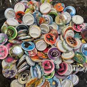 Puede incluir: Un conjunto de coloridas chapas con diversos diseños, incluyendo formas abstractas, personajes de dibujos animados y gráficos inspirados en la naturaleza. Las chapas tienen respaldos blancos con cierres de imperdible y son de varios tamaños.