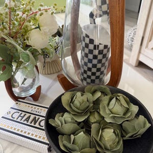 Puede incluir: Un arreglo decorativo con un jarrón de cristal transparente y un marco de madera, un portavelas a cuadros blancos y negros y un cuenco negro lleno de decoraciones verdes en forma de rosa. También hay una caja de naipes con el texto "BONNE CHANCE".