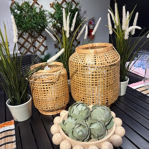 Puede incluir: Dos faroles de bambú tejido de diferentes tamaños se encuentran sobre una mesa de madera oscura, junto a un cuenco decorativo lleno de alcachofas. También hay plantas verdes altas con copas blancas y plumosas y pequeñas macetas blancas.