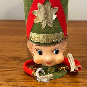 May include: A decorative elf ornament featuring a tall green felt hat with red felt trim and a gold metallic star accent. The elf has a molded plastic face with blue eyes, rosy cheeks, and a red mouth. A red felt collar with white trim surrounds the base.