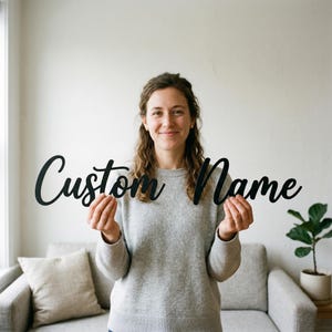 Könnte beinhalten: Ein schwarzes, maßgefertigtes Schild mit den Worten "Custom Name" in einer kursiven Schriftart. Das Schild wird von einer Person in einem grauen Pullover gehalten. Der Hintergrund umfasst ein Sofa, ein Kissen und eine Topfpflanze.