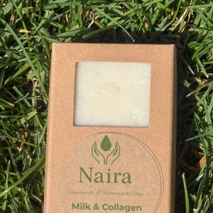Può includere: Una saponetta rettangolare è visibile attraverso una finestra in una scatola di cartone marrone. La scatola ha un adesivo circolare con il nome "Naira" e le parole "Milk & Collagen". Il sapone pesa 135 g.