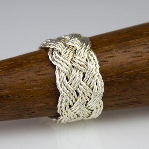 May include: A silver braided ring with a woven design. The ring is displayed on a brown wooden surface.