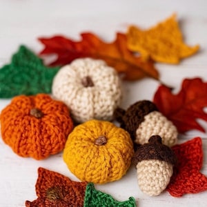 May include: A selection of handmade crochet autumn decorations, featuring pumpkins, acorns, and leaves. The pumpkins are in orange, yellow, and cream hues, with red, orange, and green leaves. The acorns have brown caps and cream-coloured bodies.