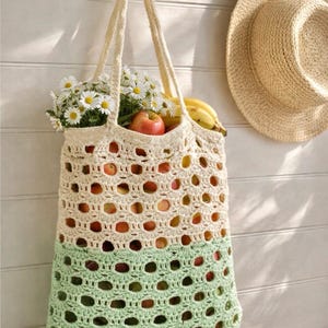 Puede incluir: Un bolso tote de crochet crema y verde claro cuelga de un gancho de pared, lleno de manzanas, plátanos y margaritas. Un sombrero de paja cuelga de un gancho cercano. El diseño de tejido abierto del bolso permite ver el contenido.