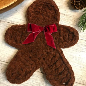 May include: A brown, plush gingerbread man-shaped item with a red velvet bow. The item has a textured surface and is likely a decorative piece or a holiday-themed item. The background includes a wooden bowl, pine needles, and a pine cone.