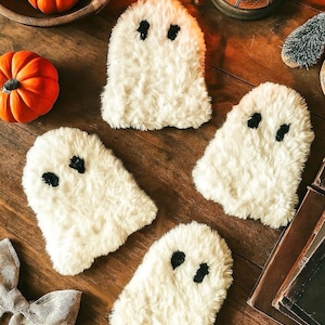 May include: Four white, fluffy ghost-shaped coasters with black eye details are arranged on a wooden surface. The Halloween-themed coasters are surrounded by miniature pumpkins and a decorative bow.