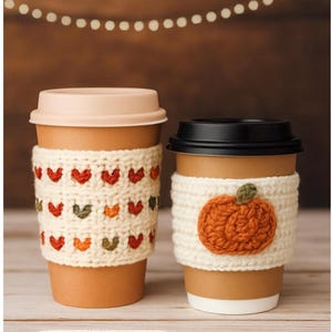 Puede incluir: Dos tazas de café con mangas de ganchillo color crema. Una manga tiene un patrón de corazones rojos, naranjas y verdes. La otra tiene una aplicación de calabaza naranja. El texto "2 in 1 Pattern!" está en la parte inferior.