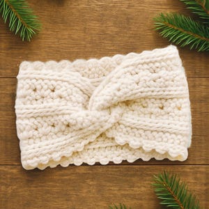 May include: Cream-colored, chunky knit headband with a twisted design. The headband has a scalloped edge and is made of a thick, textured yarn. The background is a wooden surface with green pine branches.