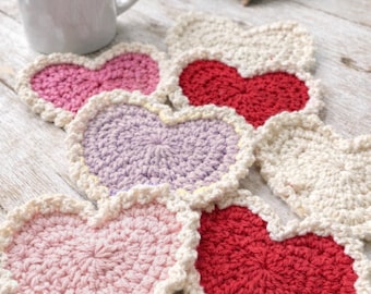 May include: A collection of handmade crochet heart-shaped coasters in various colors, including pink, red, and lavender, with a cream-colored border. A white mug with coffee sits next to the coasters. A decorative heart is in the background.