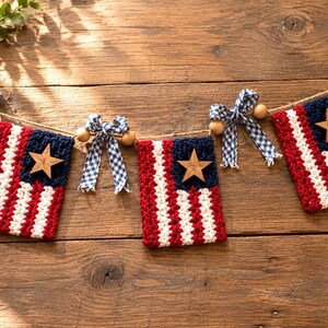 May include: A patriotic garland featuring three rectangular crocheted flags with red, white, and blue stripes. Each flag is adorned with a wooden star and separated by blue and white gingham bows and natural wooden beads on a jute twine string.