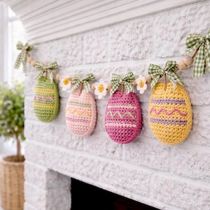 Könnte beinhalten: Eine Girlande mit gehäkelten Ostereiern in Grün, Rosa und Gelb, jedes mit einem einzigartigen Stickmuster. Die Eier sind mit grünen Vichy-Schleifen und kleinen weißen Gänseblümchen verziert, auf einer Holzperlen-Girlande aufgereiht und an einer weißen Backsteinmauer ausgestellt.