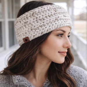 May include: A cream-colored, chunky knit headband with a small brown leather tag featuring a heart. The headband is made of thick yarn and is designed to keep the ears warm. The style is a wide band, suitable for cold weather.