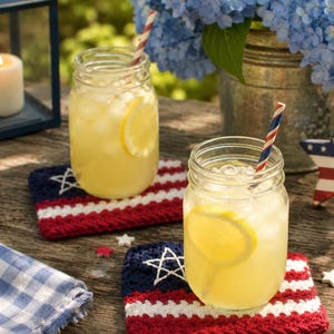 May include: Two clear glass mason jars filled with lemonade, ice, and lemon slices, each featuring a striped paper straw. The jars rest on two rectangular, crocheted coasters designed with a blue field, white stars, and red and white stripes.