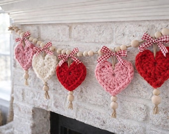 Op de afbeelding: Een handgemaakte hartjesslinger met vijf gehaakte harten in roze en rode tinten, geregen op een houten kralensnoer. Elk hart is versierd met een ruitjesstrik en een jute kwast, perfect voor Valentijnsdagdecoratie.