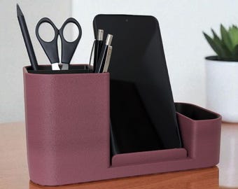 May include: A burgundy desk organizer with compartments for pens, scissors, and a smartphone. The organizer sits on a wooden desk, with small potted plants in the background. The desk organizer is a stylish way to keep your desk tidy.