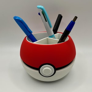 Könnte beinhalten: Ein rot-weißer Schreibtisch-Organizer in Form eines Pokéballs, mit einem schwarzen Band und einem weißen Knopf. Der Organizer enthält blaue, schwarze und türkisfarbene Stifte und Marker.