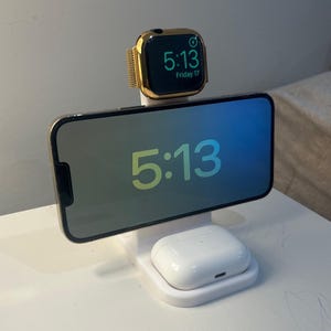 Könnte beinhalten: Eine weiße Ladestation mit einer goldenen Apple Watch, die 5:13 Freitag 17 anzeigt, einem Telefon mit 5:13 und einem weißen AirPods-Etui. Telefon und Uhr sind goldfarben.
