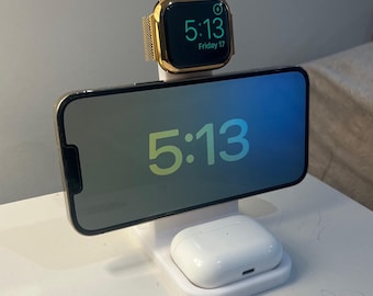 May include: A white charging station with a gold Apple Watch displaying 5:13 Friday 17, a phone showing 5:13, and a white AirPods case. The phone and watch are gold-colored.