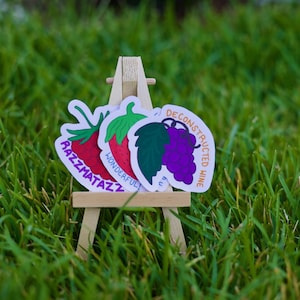May include: Three colorful stickers on a miniature wooden easel, set against a backdrop of green grass. The stickers feature illustrations of a strawberry, a grape cluster, and a strawberry with the text "Razzmatazz", "Wonderful!", and "Deconstructed Wine".