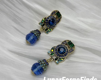 May include: A pair of ornate blue and gold earrings. Each earring features a large, round, deep blue crystal at the center, surrounded by smaller blue and green crystals. A dangling blue crystal bead hangs below. The earrings have a vintage aesthetic.