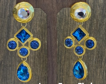 May include: A pair of gold-toned earrings with a geometric design. Each earring features a large, round top with a clear, faceted stone, a blue square, three blue circles, and a teardrop-shaped blue stone. The earrings are displayed on a wooden stand.