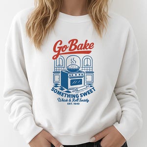 May include: White sweatshirt with a blue and red graphic that says "Go Bake Something Sweet" with an illustration of a pie and oven. The sweatshirt also says "Whisk & Roll Society Est. 1940".