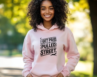 Puede incluir: Sudadera con capucha rosa claro con bolsillo delantero y capucha con cordón. El texto negro en la parte delantera dice "I GET PAID TO BE PULLED DOWN THE STREET" con un pequeño gráfico de perro. La sudadera está hecha de un material suave.
