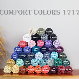 May include: A display of rolled t-shirts in various colors, arranged in a pyramid shape. The image includes the text "COMFORT COLORS 1717" at the top. Colors include black, blue, green, and red.