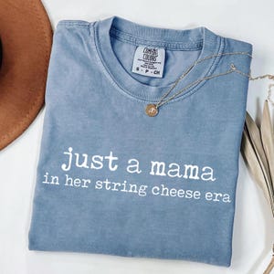 May include: A blue t-shirt with the text "just a mama in her string cheese era" in white. A gold necklace with a pendant is draped across the shirt. The shirt is folded on a white surface.