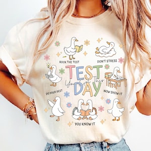 May include: Cream-colored t-shirt with a colorful graphic featuring cartoon ducks and the text "TEST DAY". The ducks are depicted in various poses with encouraging phrases like "Rock the Test" and "Do Your Best".