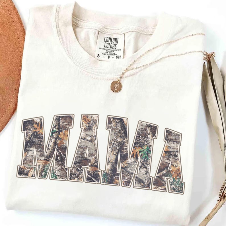 May include: Cream-colored t-shirt with the word "MAMA" in a camouflage design. A gold necklace with a round pendant rests on the shirt. The shirt has a Comfort Colors tag.