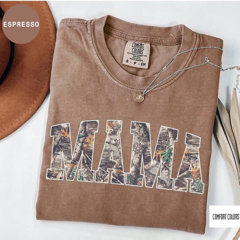 May include: Espresso-colored Comfort Colors t-shirt with the word "MAMA" in a camouflage design. A silver necklace with a pendant rests on the shirt. The shirt is a small size.