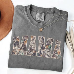 May include: A gray Comfort Colors t-shirt with the word "MAMA" in a camouflage pattern. A silver necklace with a pendant is draped across the shirt. A brown hat is visible in the upper left corner.