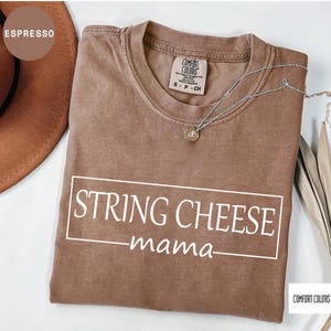 May include: Espresso-colored t-shirt with the text "STRING CHEESE mama" in a white rectangular box. The shirt has a crew neck and short sleeves. A silver necklace with a pendant is draped on the shirt.