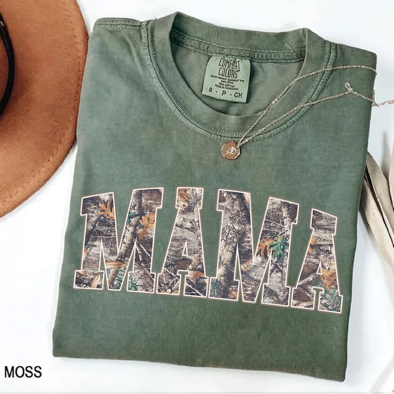 May include: Moss green t-shirt with the word "MAMA" in a camouflage pattern. The shirt has a crew neck and short sleeves. A silver necklace with a small pendant is draped on the shirt.