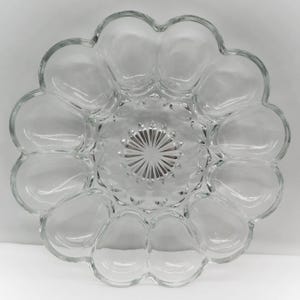 May include: A clear glass egg serving platter featuring a scalloped outer edge with twelve individual oval-shaped indentations arranged in a circular pattern around a central starburst design with radiating etched lines.