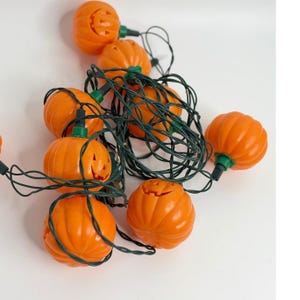May include: A string of decorative lights featuring multiple orange plastic pumpkin-shaped covers. Each pumpkin has carved facial features and is attached to a dark green electrical cord with small green plastic connectors.
