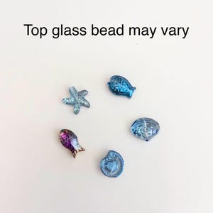 Peut inclure: Cinq petites perles en verre en forme de cr&eacute;atures marines, dont une &eacute;toile de mer, deux poissons et deux coquillages. Les perles sont principalement bleues, avec un poisson aux tons iris&eacute;s violet et or. Le texte Top glass bead may vary est visible.