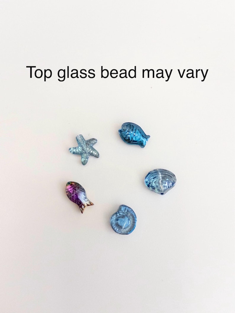 Peut inclure: Cinq petites perles en verre en forme de cr&eacute;atures marines, dont une &eacute;toile de mer, deux poissons et deux coquillages. Les perles sont principalement bleues, avec un poisson aux tons iris&eacute;s violet et or. Le texte Top glass bead may vary est visible.