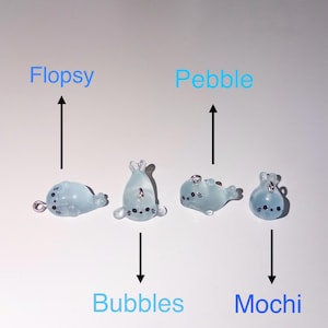 Peut inclure: Quatre breloques en forme de phoque bleu clair translucide avec des yeux et des moustaches peints en noir. Chaque breloque comporte une petite boucle m&eacute;tallique sur le dessus ou sur le c&ocirc;t&eacute;. Les noms Flopsy, Bubbles, Pebble et Mochi sont indiqu&eacute;s par des fl&egrave;ches noires pointant vers chaque figurine.