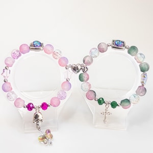May include: Two beaded bracelets with a variety of colorful beads in pink, purple, and green hues. One bracelet features a skull charm and a butterfly charm, while the other has a cross charm. Both bracelets have a heart-shaped clasp.