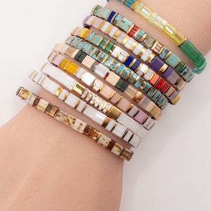 May include: A collection of colorful beaded bracelets. The bracelets feature a variety of colors including turquoise, white, gold, and red. Each bracelet is made of small, rectangular beads and is accented with gold-colored metal. The bracelets are stacked on a wrist.