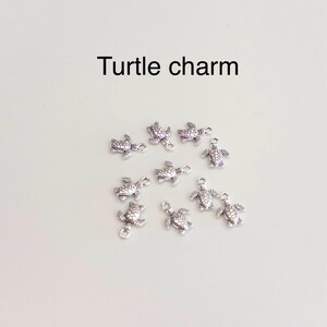Peut inclure: Un ensemble de neuf petites breloques en m&eacute;tal argent&eacute; en forme de tortue, chacune pr&eacute;sentant des motifs de carapace d&eacute;taill&eacute;s et une petite boucle sur le dessus. Le texte 'Turtle charm' est imprim&eacute; en noir au-dessus du groupe.